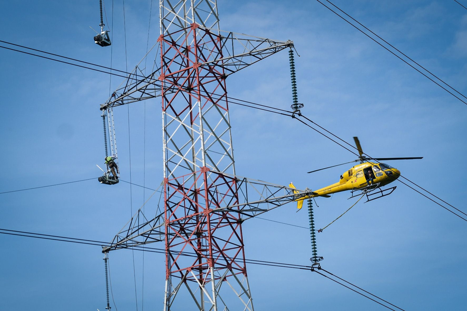 De man op het laddertje hoog in de mast legt de trekkoord over de wieltjes terwijl zijn collega in de helikopter nauwgezet toekijkt.